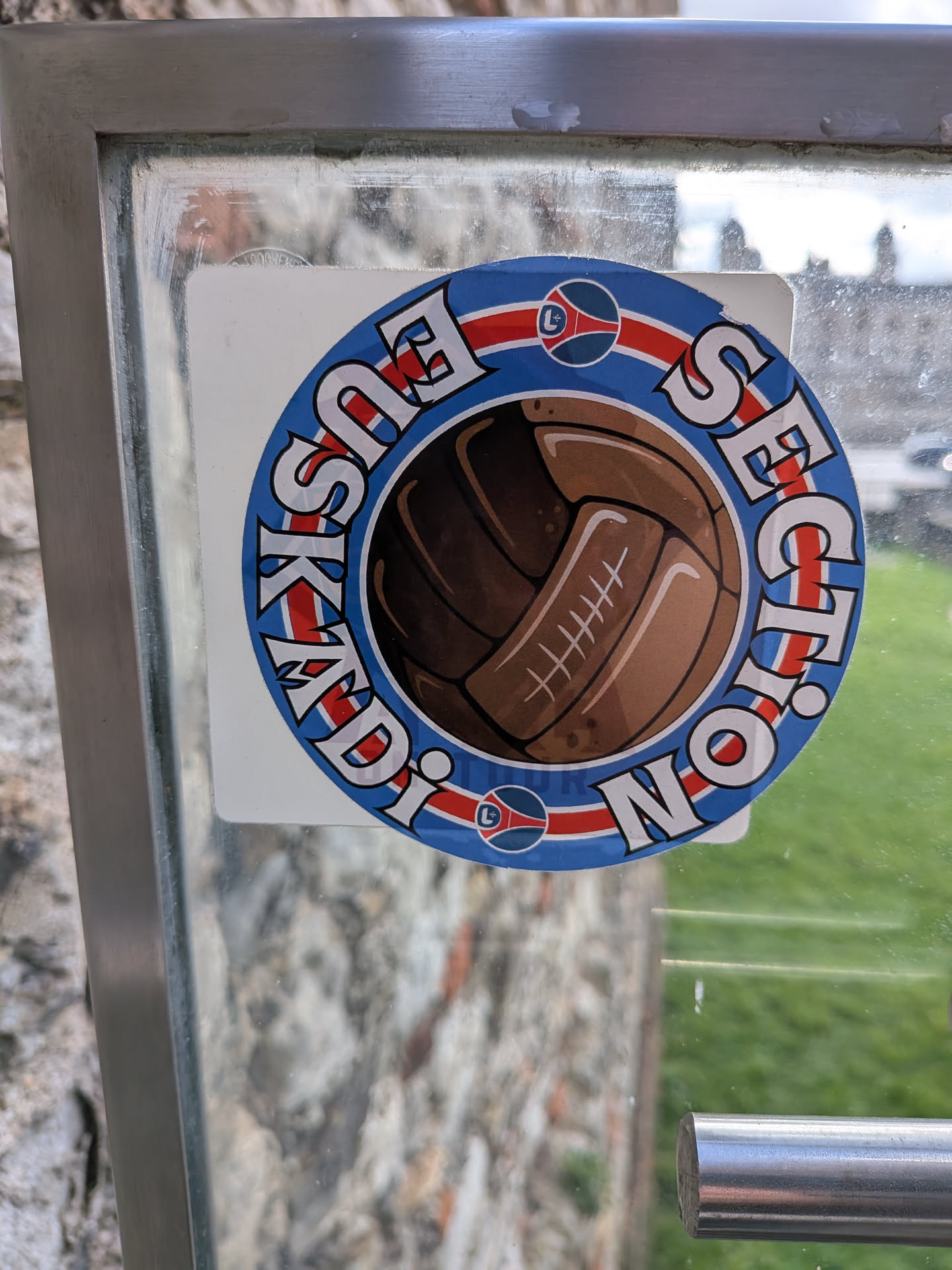 A circular sticker featuring a vintage brown leather football at its center. The football is detailed with white stitching. Surrounding the football is a blue ring with white, block-style text outlined in black. A horizontal red and white striped band runs through the middle of the blue ring, passing behind the football. On this band, at the 3 and 9 o'clock positions, are small circular logos with a stylized 'L' in red and blue. The sticker is applied to a glass surface, showing some minor wear and slight peeling at the top right.