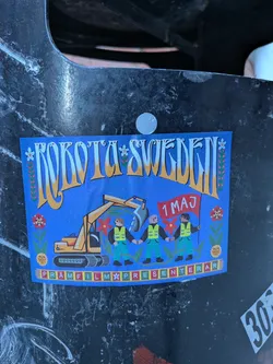A rectangular sticker with a bright blue background, rendered in a folk art style. At the top, stylized yellow-orange text with white highlights reads 'ROKOTA SWEDEN', adorned with small white flowers and green leafy vines. The central illustration depicts a yellow excavator on the left, surrounded by green foliage and red flowers. To the right, three figures in green pants and yellow safety vests hold hands, forming a chain. The figure on the far right holds a red flag with '1 MAJ' (1st May) written in white. The ground beneath the figures is green with more red flowers and a stylized sun face. Along the bottom edge, a decorative border of small colored squares (red, green, yellow, white) contains the text 'PRAMFILM PRESENTERAR'. The overall aesthetic is colorful, detailed, and conveys a message of labor and community.