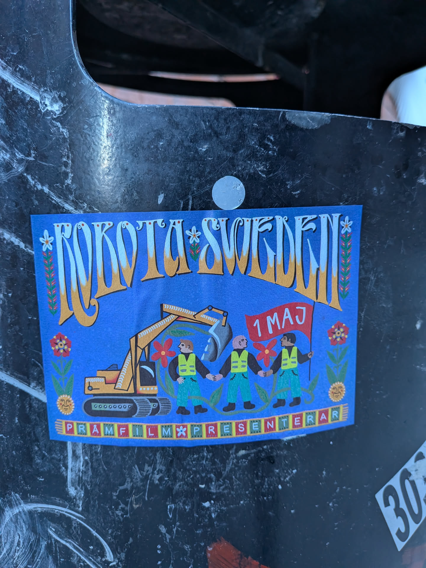 A rectangular sticker with a bright blue background, rendered in a folk art style. At the top, stylized yellow-orange text with white highlights reads 'ROKOTA SWEDEN', adorned with small white flowers and green leafy vines. The central illustration depicts a yellow excavator on the left, surrounded by green foliage and red flowers. To the right, three figures in green pants and yellow safety vests hold hands, forming a chain. The figure on the far right holds a red flag with '1 MAJ' (1st May) written in white. The ground beneath the figures is green with more red flowers and a stylized sun face. Along the bottom edge, a decorative border of small colored squares (red, green, yellow, white) contains the text 'PRAMFILM PRESENTERAR'. The overall aesthetic is colorful, detailed, and conveys a message of labor and community.