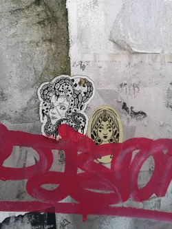 The image features two distinct, hand-drawn style stickers applied to a textured, grey and white wall, partially covered by vibrant red graffiti. The left sticker, larger and irregularly shaped, depicts a woman's head and shoulders in profile, looking to the left. Her hair is intricately detailed with snakes and swirling patterns, reminiscent of Medusa, rendered in black and white line art with subtle shading. Red paint drips from the bottom edge of this sticker, blending with the graffiti below. The right sticker, smaller and oval-shaped, presents a woman's face looking directly forward. She has stylized eyes with heart-shaped pupils and an elaborate, symmetrical headdress or hairstyle. This sticker is also executed in black and white line art. A band of red paint from the graffiti covers the lower part of her face, partially obscuring some text.