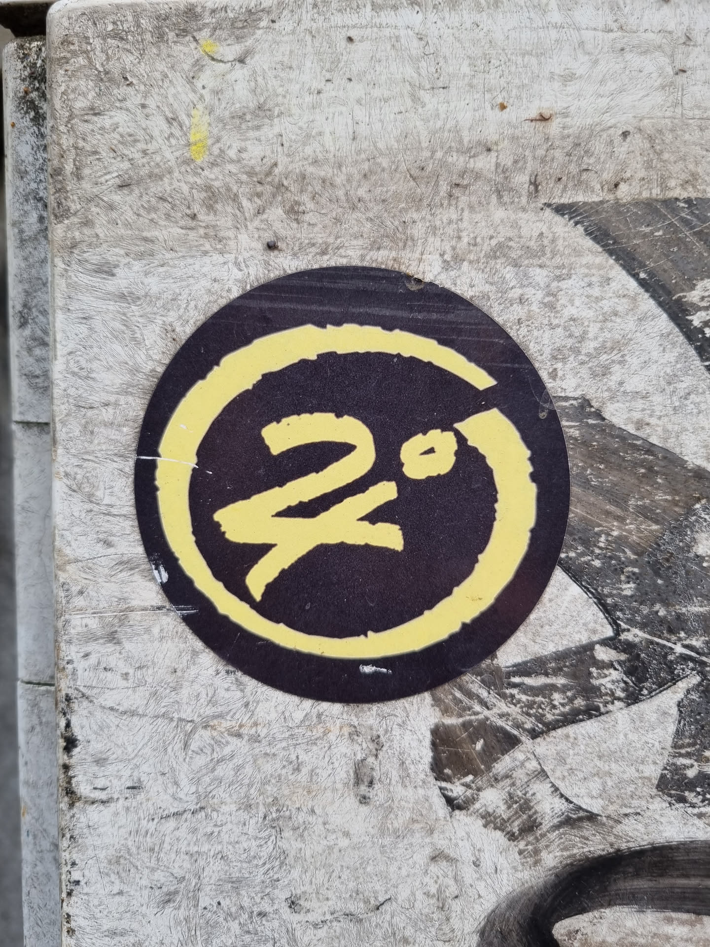 A round, dark-colored sticker with a yellow circle and stylized characters in the center. The sticker appears to be affixed to a weathered surface. The central characters are not easily identifiable.