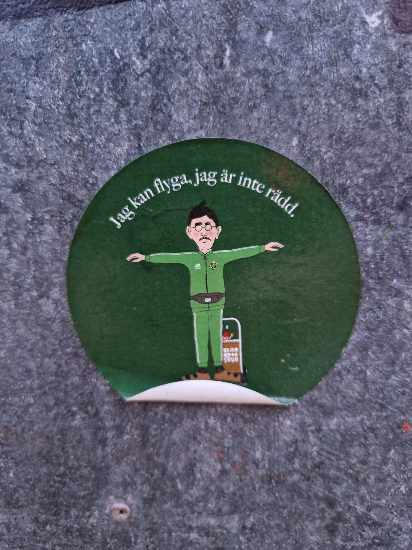 A round green sticker with an illustration of a man with his arms outstretched, as if flying. The text around the illustration says: "Jag kan flyga, jag är inte rädd" which translates to  "I can fly, I'm not afraid" in Swedish. The man is wearing green clothes and a belt bag.