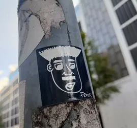 This black and white sticker depicts a cartoon-like face with a distinctive pompadour hairstyle. The face is simply drawn with bold outlines. The sticker is affixed to a weathered metal pole.