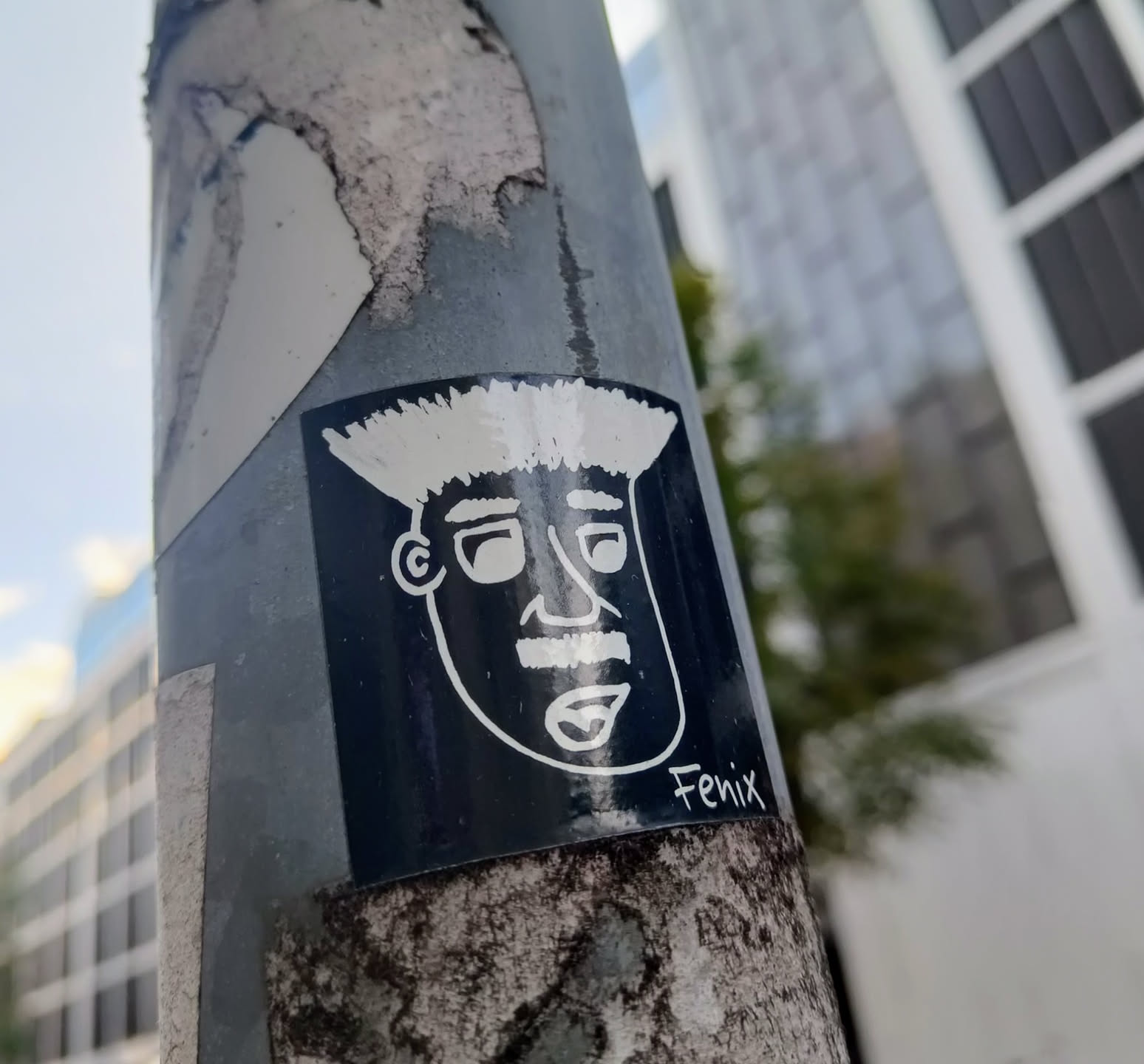 This black and white sticker depicts a cartoon-like face with a distinctive pompadour hairstyle. The face is simply drawn with bold outlines. The sticker is affixed to a weathered metal pole.