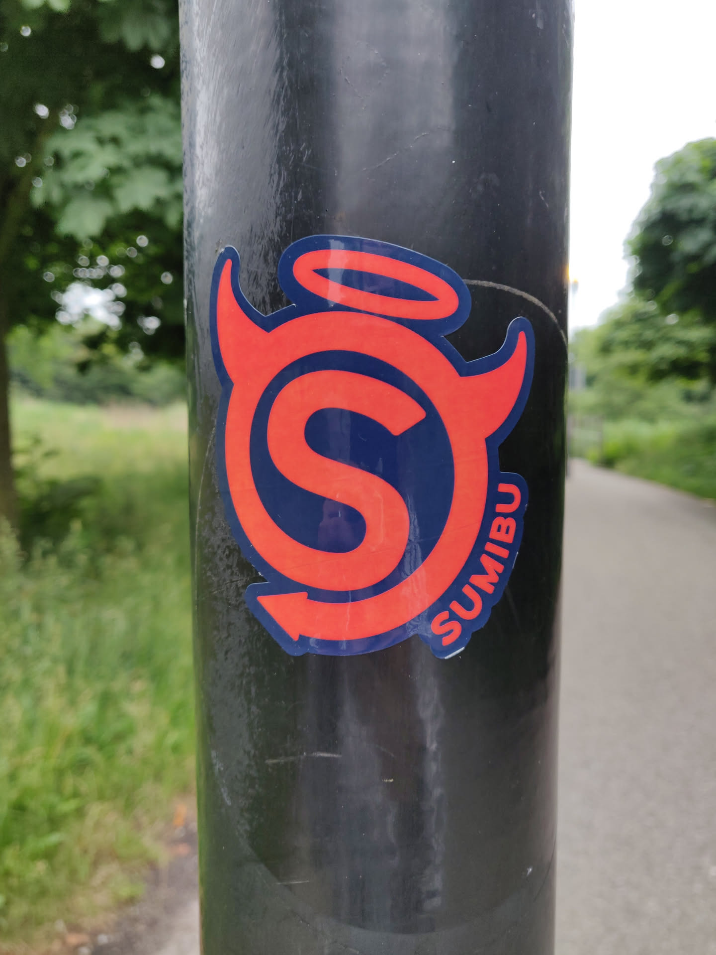 A sticker featuring a stylized 'S' logo with devil horns and a halo. The word SUMIBU is written along the bottom curve of the design. The sticker is predominantly red and dark blue, applied to a black pole. 