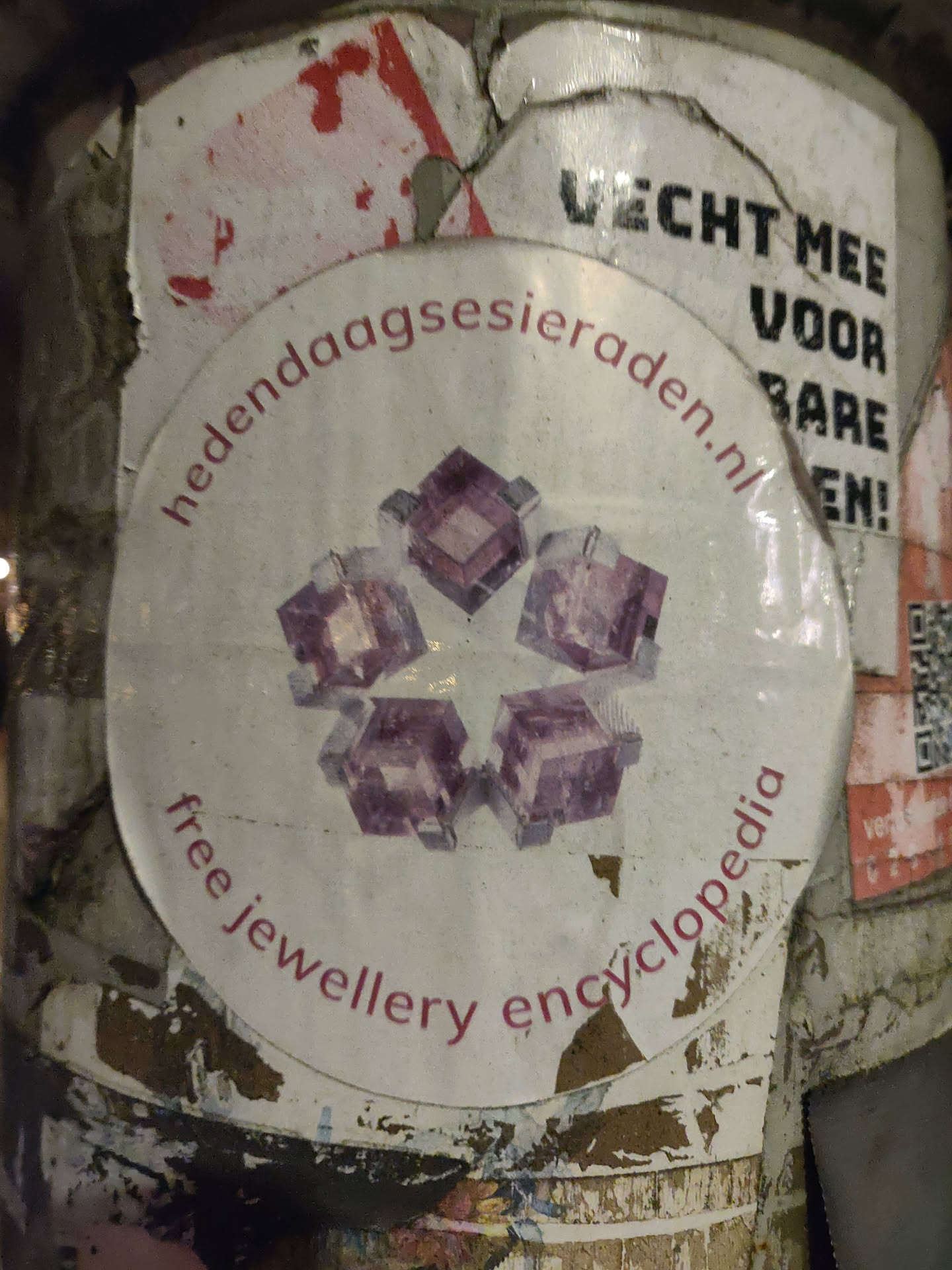 A round sticker with a pink design of what looks like crystals or gemstones arranged in a star shape. The text on the sticker says "Free Jewellery Encyclopedia" and also shows a website address: hedendaagssieraden.nl
