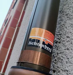 A retro-style sticker featuring an illustration of the Lichtenberg skyline with a sunset backdrop. The text 'Lichtenberg' is prominently displayed.  It's stuck on a dark-green downspout with copper accents.
