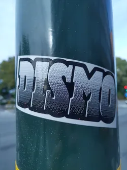 A black and white sticker with the word DISMO in a stylized halftone font. The sticker is applied to a dark green pole.