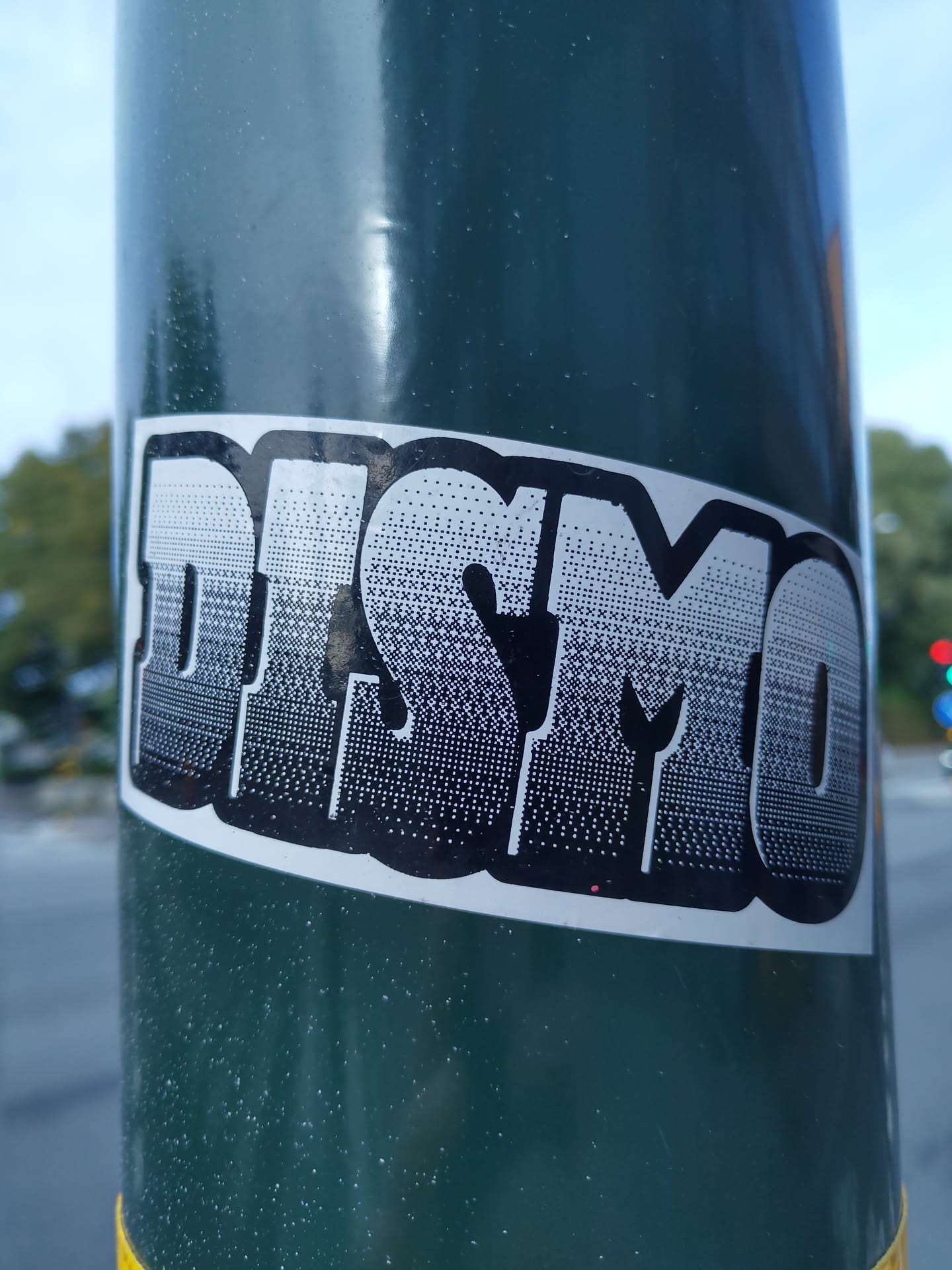 A black and white sticker with the word DISMO in a stylized halftone font. The sticker is applied to a dark green pole.