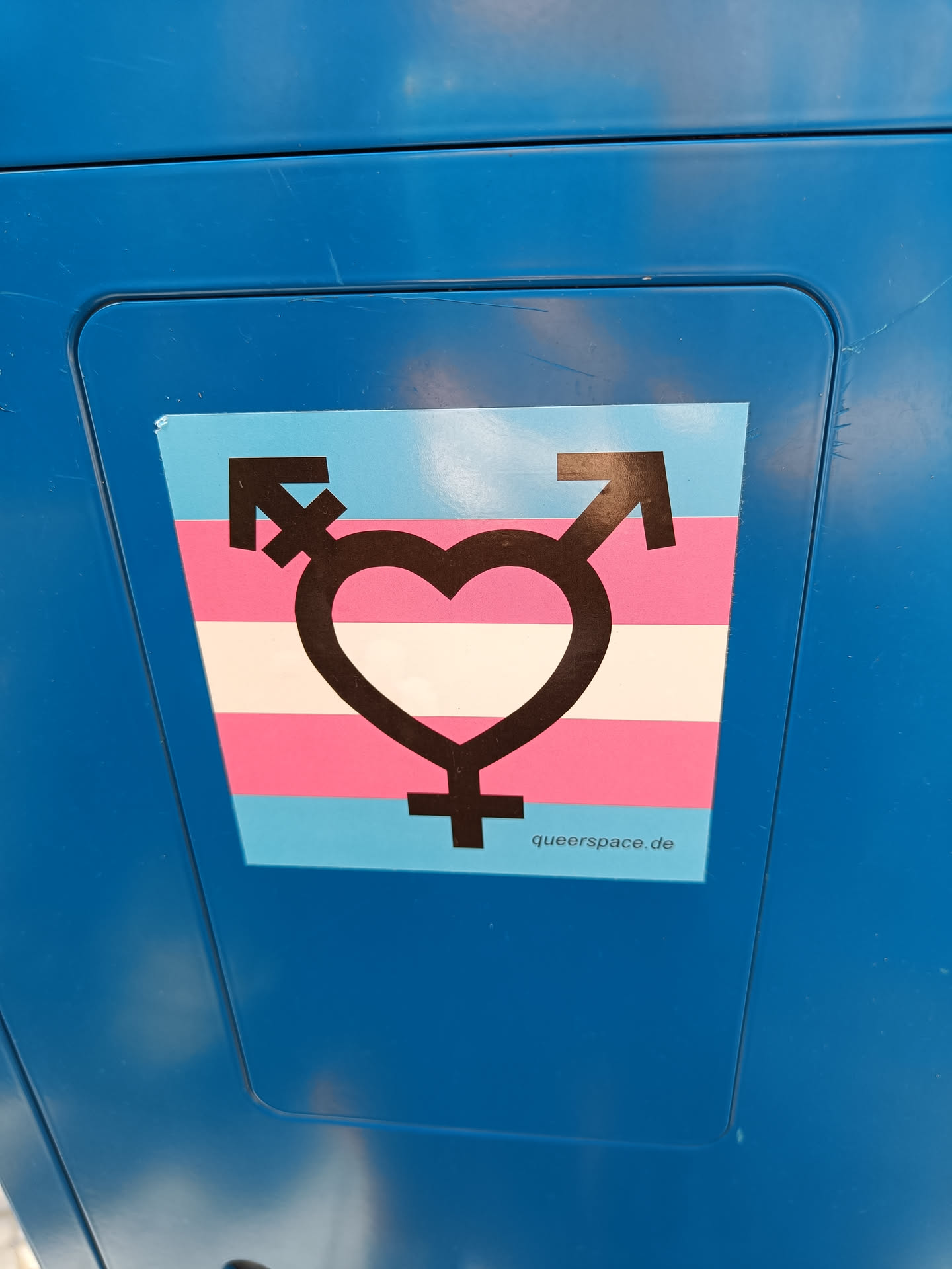 A sticker featuring a heart symbol combined with transgender symbols, set against the transgender flag colors.  The website queerspace.de is visible at the bottom.