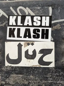 The image features two stickers stacked on top of each other on a rough, textured surface. The top two stickers are rectangular with the word "KLASH" printed twice in bold, white letters against a black background. The bottom sticker is white with a black, stylized design that resembles Arabic text. The surface behind the stickers appears to be weathered and marked with various stains or graffiti.