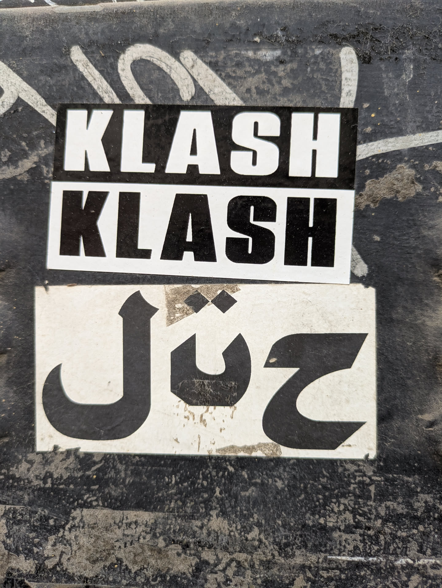The image features two stickers stacked on top of each other on a rough, textured surface. The top two stickers are rectangular with the word "KLASH" printed twice in bold, white letters against a black background. The bottom sticker is white with a black, stylized design that resembles Arabic text. The surface behind the stickers appears to be weathered and marked with various stains or graffiti.