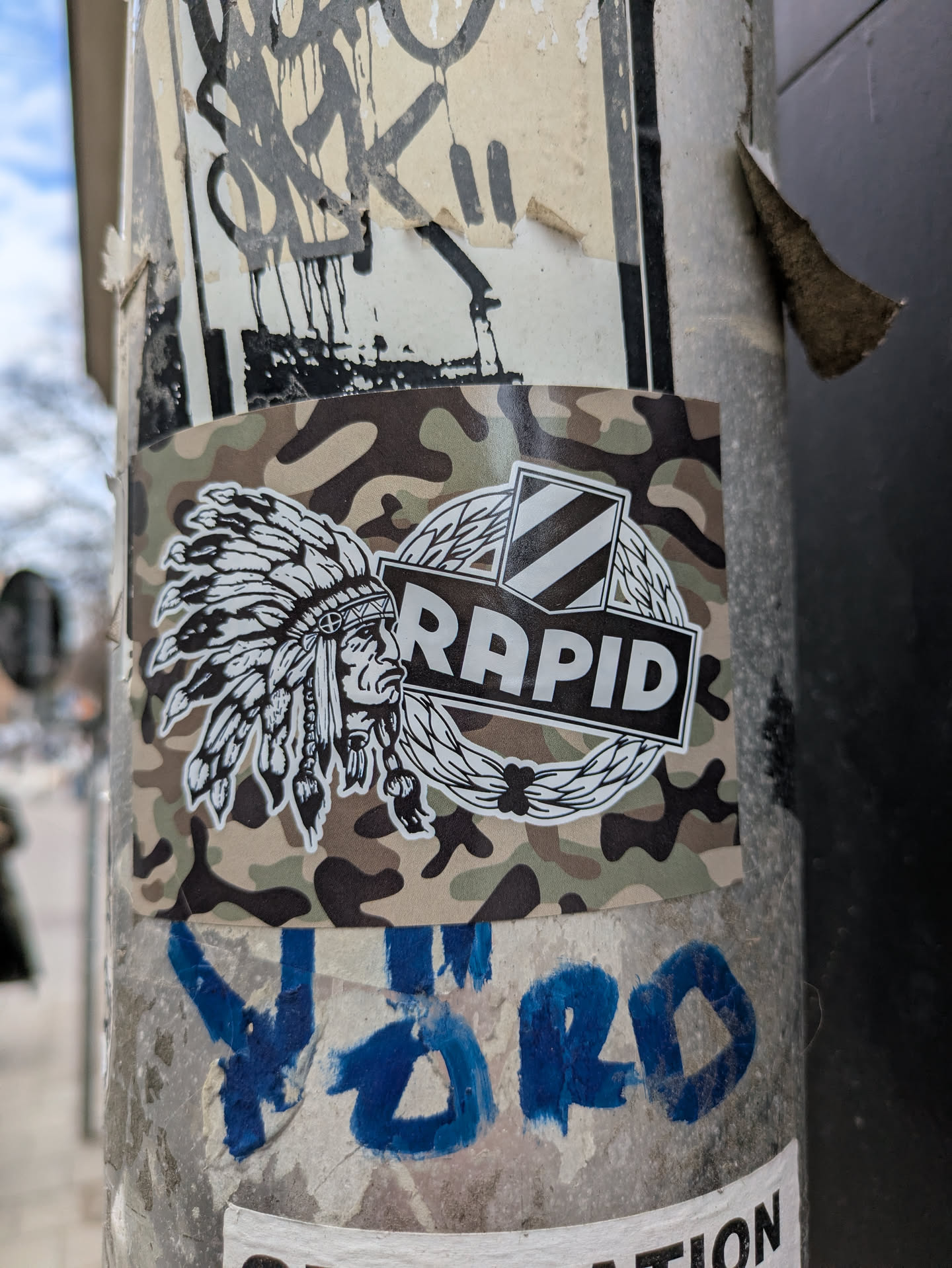 The sticker features a detailed depiction of a Native American chief wearing a traditional headdress, positioned next to the word "Rapid" enclosed in a circular emblem, against a camouflage background. The design gives a unique blend of cultural symbolism and modern design elements.