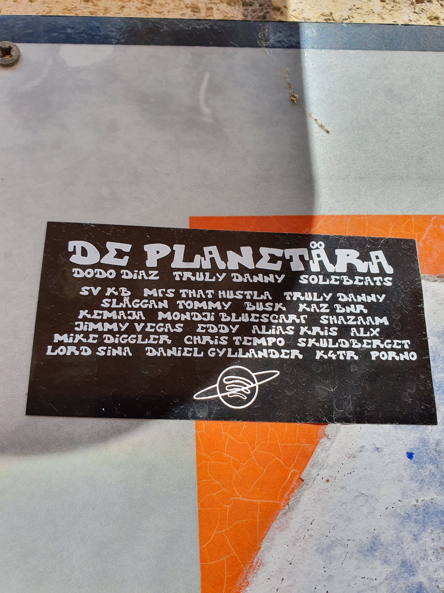 A black and white sticker with the text "DE PLANETÄRA" at the top and a list of names underneath. There is a small symbol at the bottom, which might be a logo. The sticker seems to be related to music or artists.
