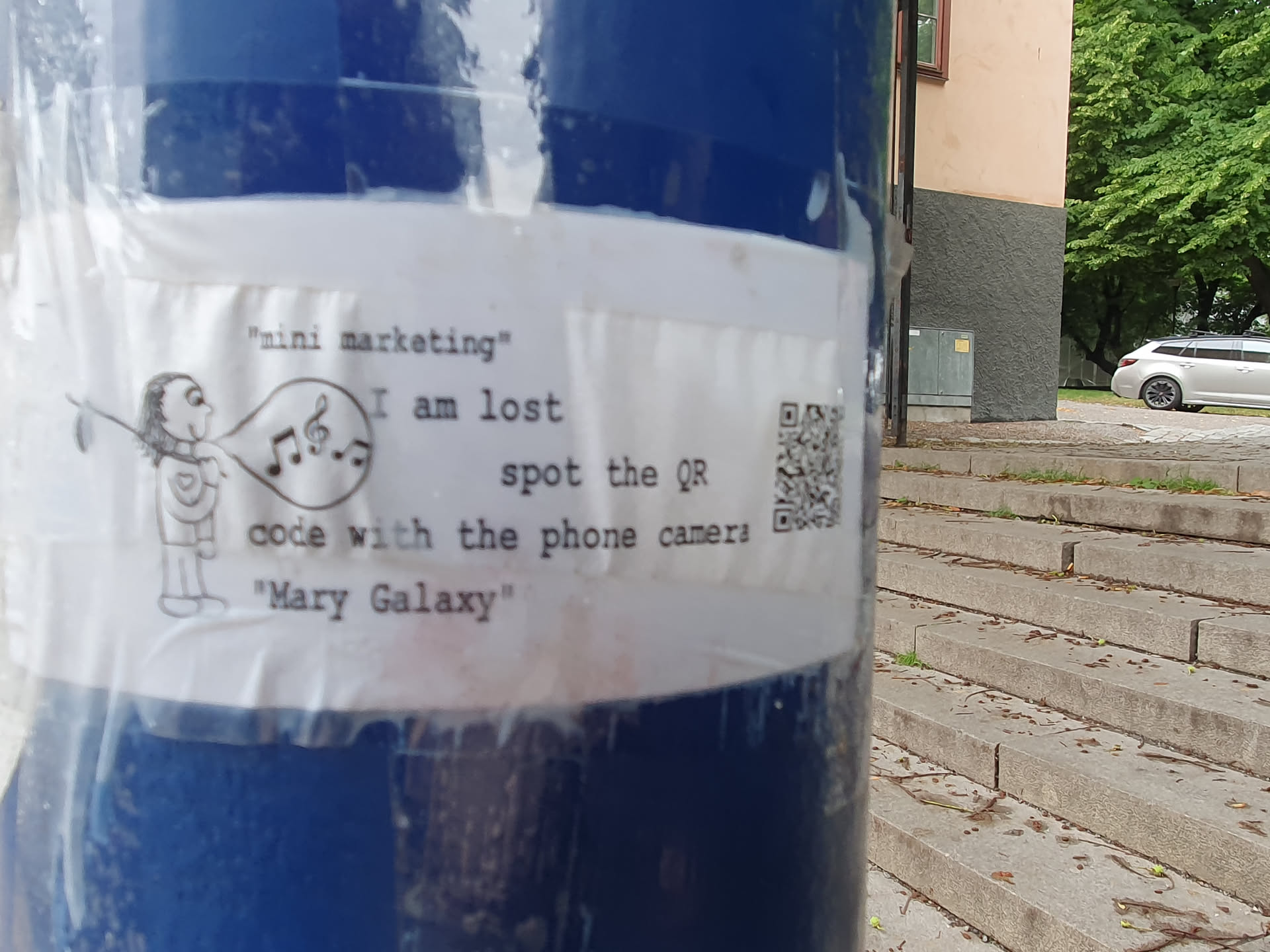 A sticker with a drawing of a person playing a flute and a QR code. The text on the sticker says "mini marketing", "I am lost", "spot the QR code with the phone camera", and "Mary Galaxy".