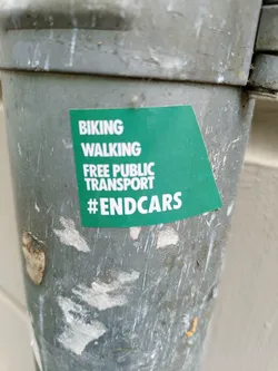 A dark-green square sticker is affixed to a weathered, gray metal pole. The sticker displays the text 'BIKING WALKING FREE PUBLIC TRANSPORT' stacked vertically, followed by the hashtag '#ENDCARS' in a slightly larger font size.  The sticker shows some minor wear and tear along its edges.
