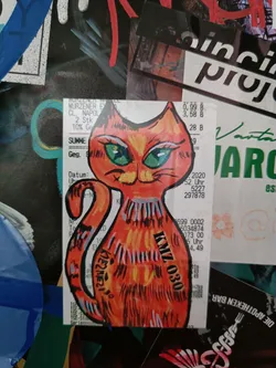 A hand-drawn sticker of an orange cat with big green eyes. The sticker is affixed to a receipt. The cat is facing forward and its body is slightly angled to the left. The cat's tail is curved. The sticker has some black markings which appear to be a signature or possibly a tag.