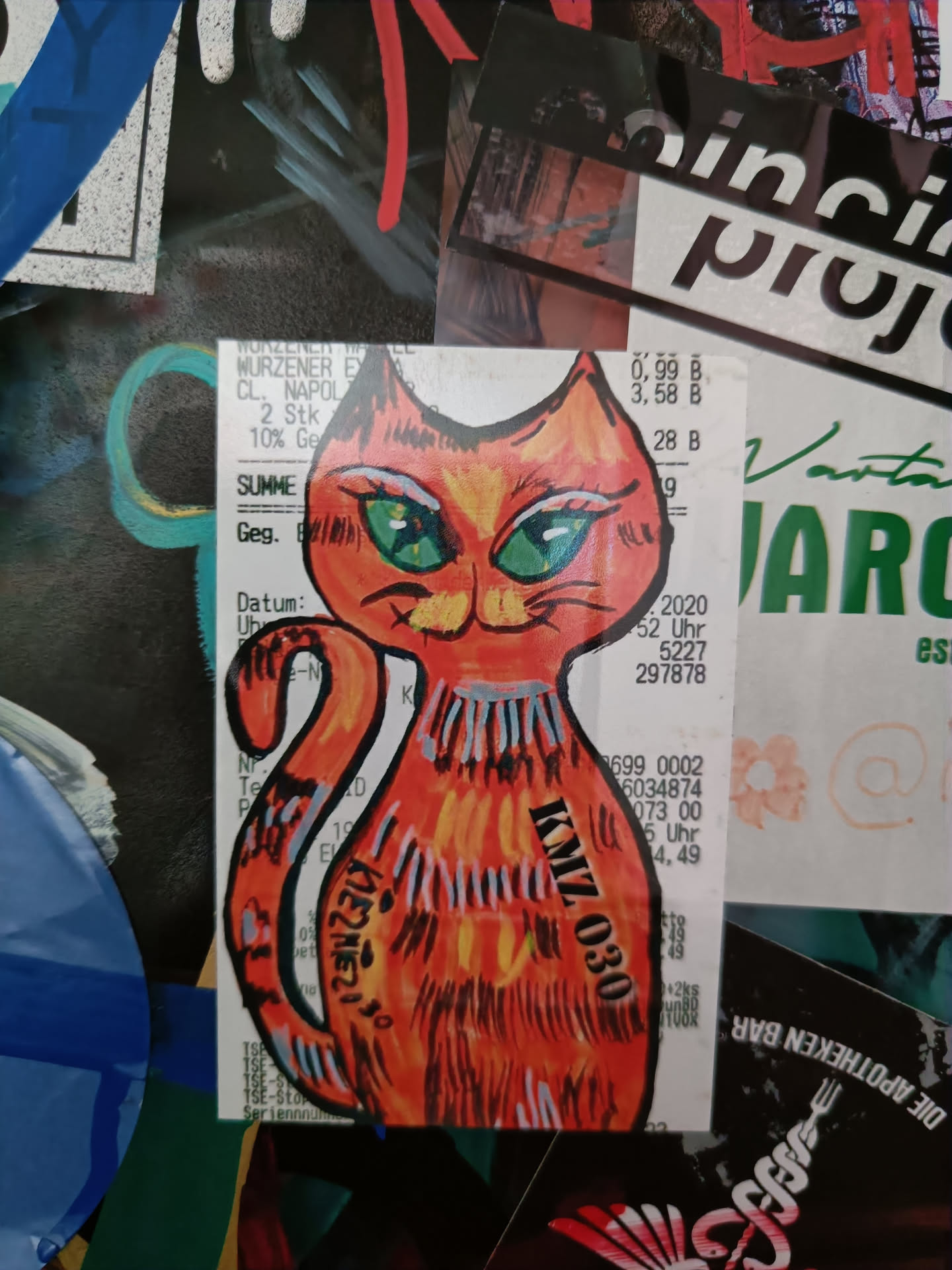 A hand-drawn sticker of an orange cat with big green eyes. The sticker is affixed to a receipt. The cat is facing forward and its body is slightly angled to the left. The cat's tail is curved. The sticker has some black markings which appear to be a signature or possibly a tag.