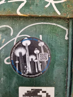 Round sticker with a black and white illustration of tall, slender towers resembling mushrooms or futuristic architecture at night. The text '@understyle.shop' is repeated around the circular sticker. A QR code is present. 