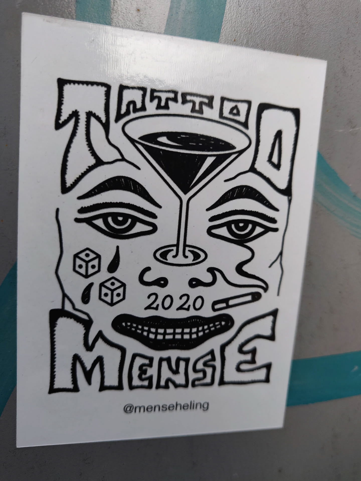 Black and white sticker with a stylized face, a martini glass, dice, and a cigarette. The text 'Tattoo MenSe' and '2020' are visible.  The artist's Instagram handle is included at the bottom.