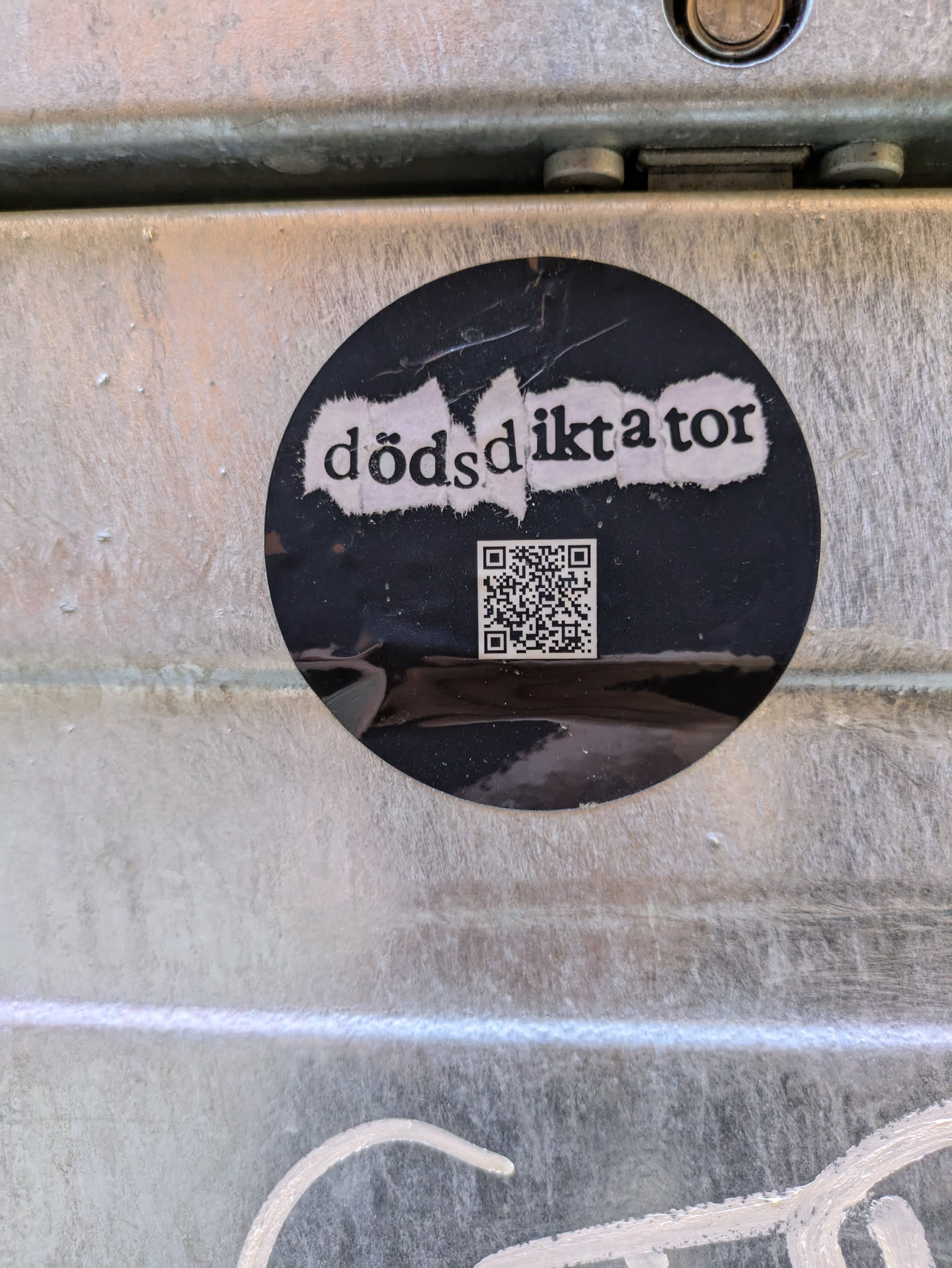 A black round sticker is affixed to a metallic surface. The sticker has a white text "dödSdiktator" and a QR code below it. The QR code is scannable.