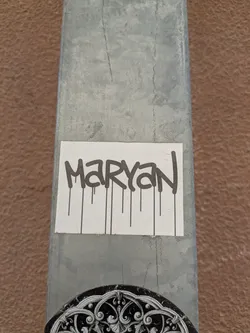 A sticker is adhered to a gray metal pole. The sticker has the name "MARYAN" written in black, dripping style letters on a white background. Another sticker featuring a decorative black and white design is partially visible below it.