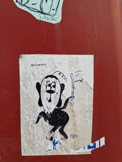 A weathered sticker featuring a unique cartoon character design by Mister Tulp. The character is depicted in black and white, holding a small flag with a heart on it. The sticker shows signs of wear and tear, adding to its character.