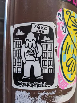 Black and white sticker with a cartoon character holding a sign that says "Zeke was here." The character is standing in front of buildings. The sticker also includes the artist's Instagram handle.