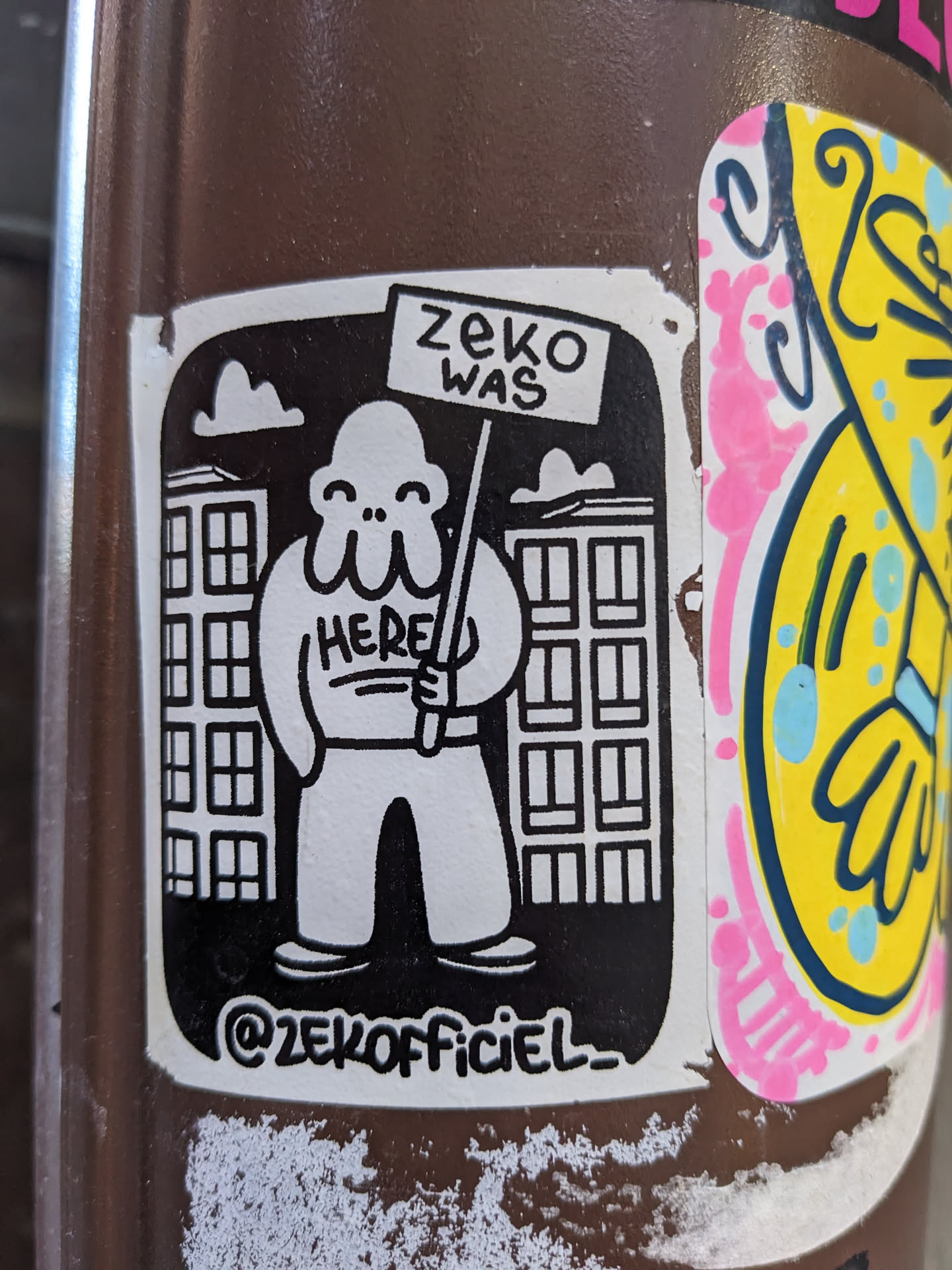 Black and white sticker with a cartoon character holding a sign that says "Zeke was here." The character is standing in front of buildings. The sticker also includes the artist's Instagram handle.