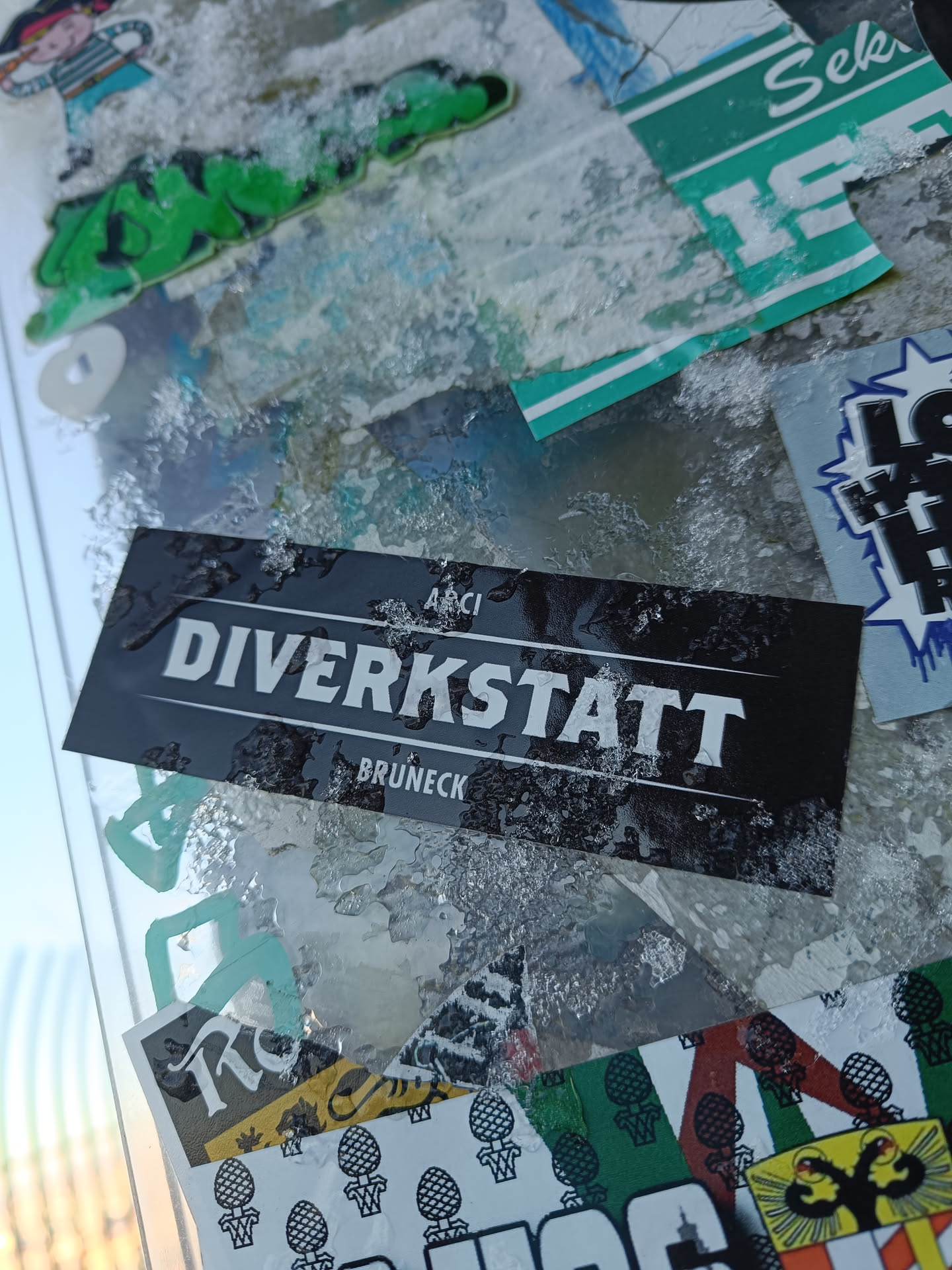 A black sticker with white text. The text says "DIVERKSTATT" with "BRUNECK" underneath it and "AACI" on the top right corner. The sticker is covered in frost or ice.