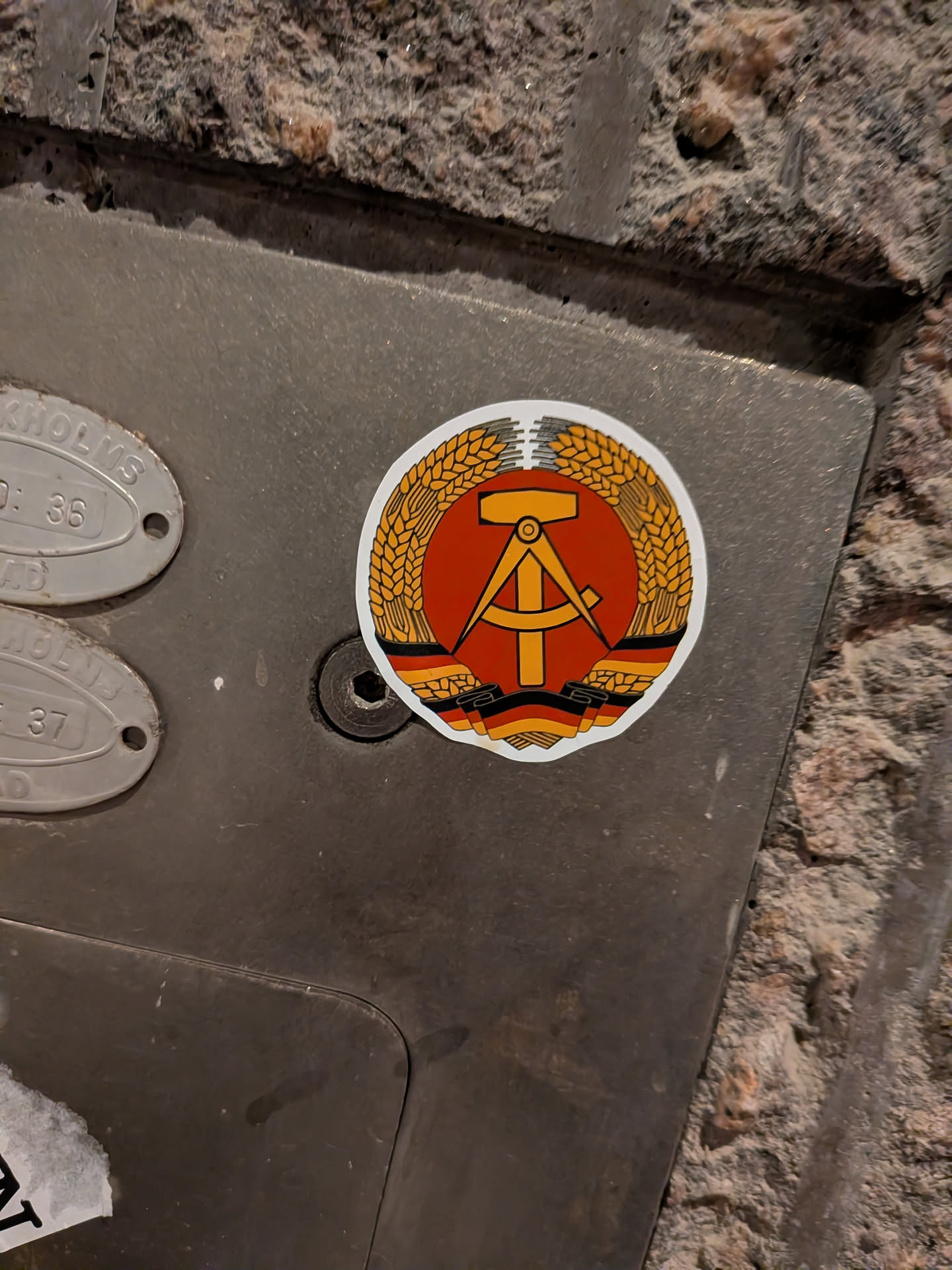 A circular sticker featuring the national emblem of the German Democratic Republic (GDR). The central design consists of a golden hammer and a golden compass, overlaid on a vibrant red circular field. This central motif is encircled by a wreath of golden rye. At the very bottom of the emblem, a ribbon in the colors of the German flag (black, red, and gold) is visible. The entire emblem is set against a clean white border, indicating it is a cut-out sticker.