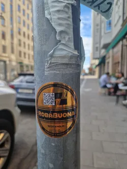 A gold-colored circular sticker featuring the text "ROBABUONA" over "STOCKHOLM" and the words "GASTRONOMI & DELIKATESSENS" surrounding the main text. It also includes a QR code. The sticker is affixed to a metallic pole on a city street.