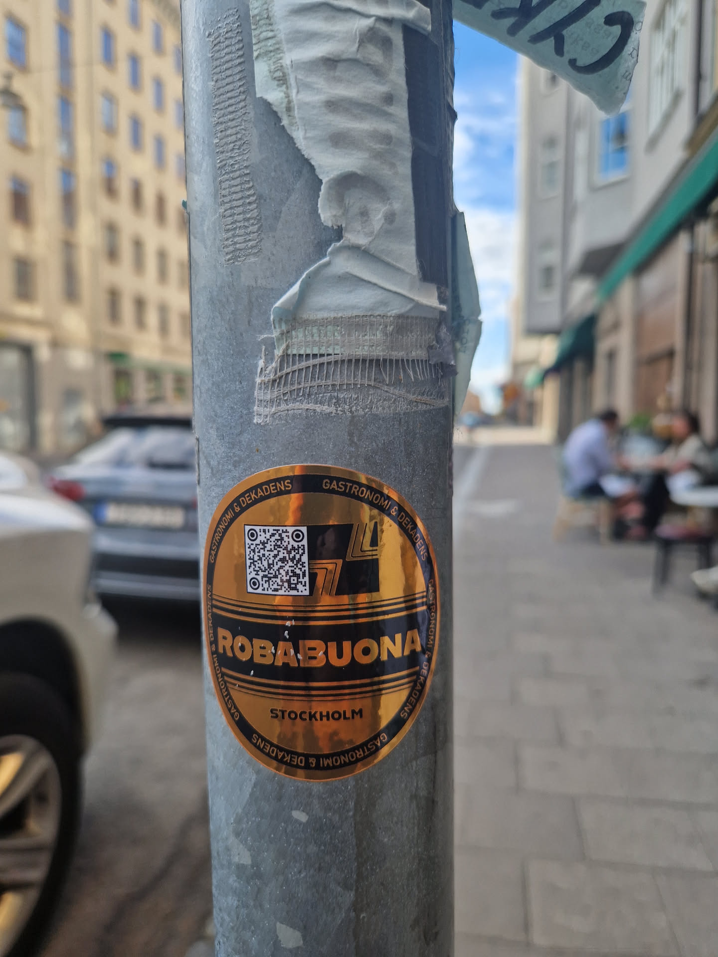 A gold-colored circular sticker featuring the text "ROBABUONA" over "STOCKHOLM" and the words "GASTRONOMI & DELIKATESSENS" surrounding the main text. It also includes a QR code. The sticker is affixed to a metallic pole on a city street.