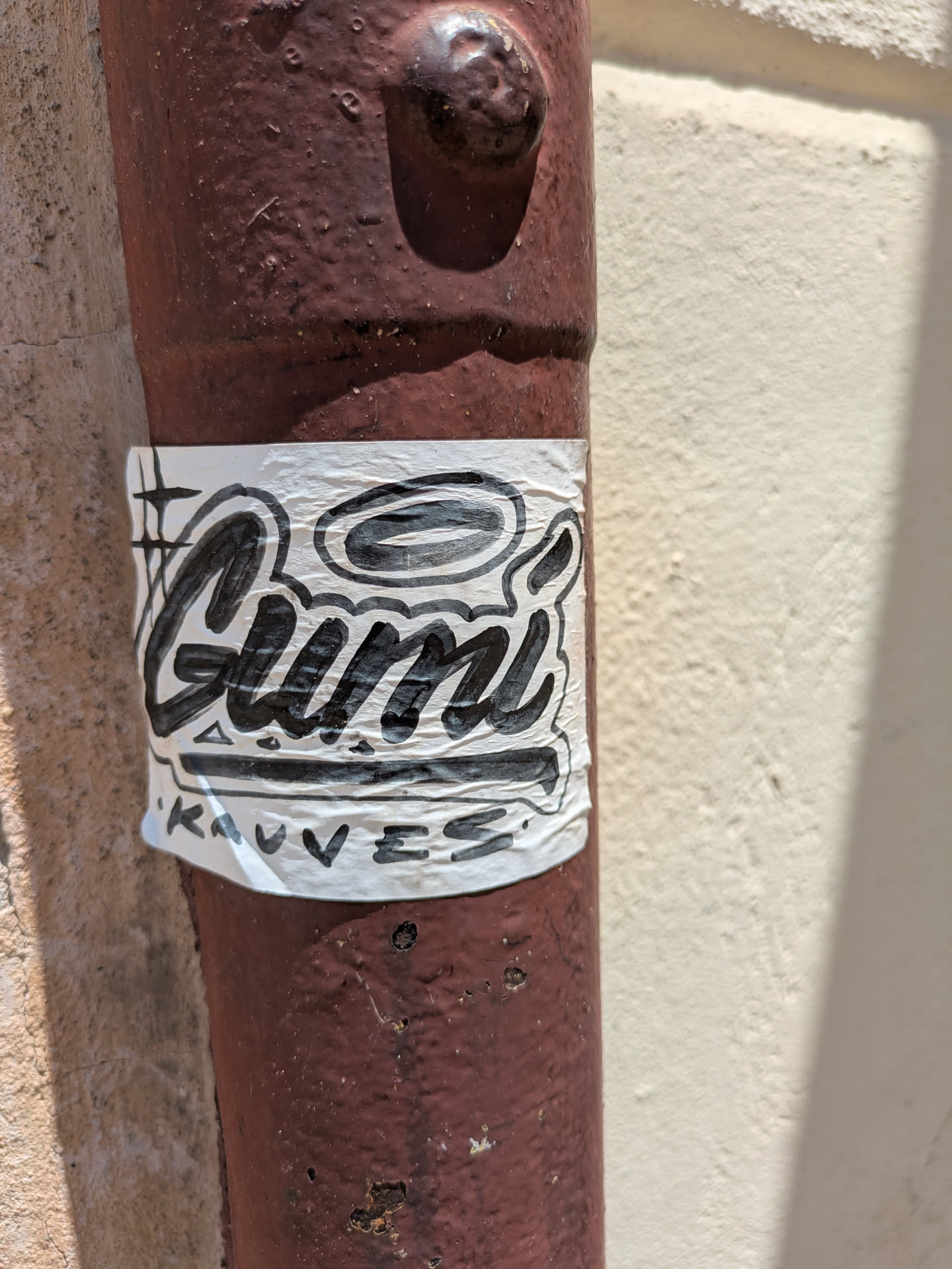 A white sticker with black, stylized graffiti lettering that reads 'Gumi' and underneath, '‘Kauves’'. The sticker is attached to a red metal pipe.
