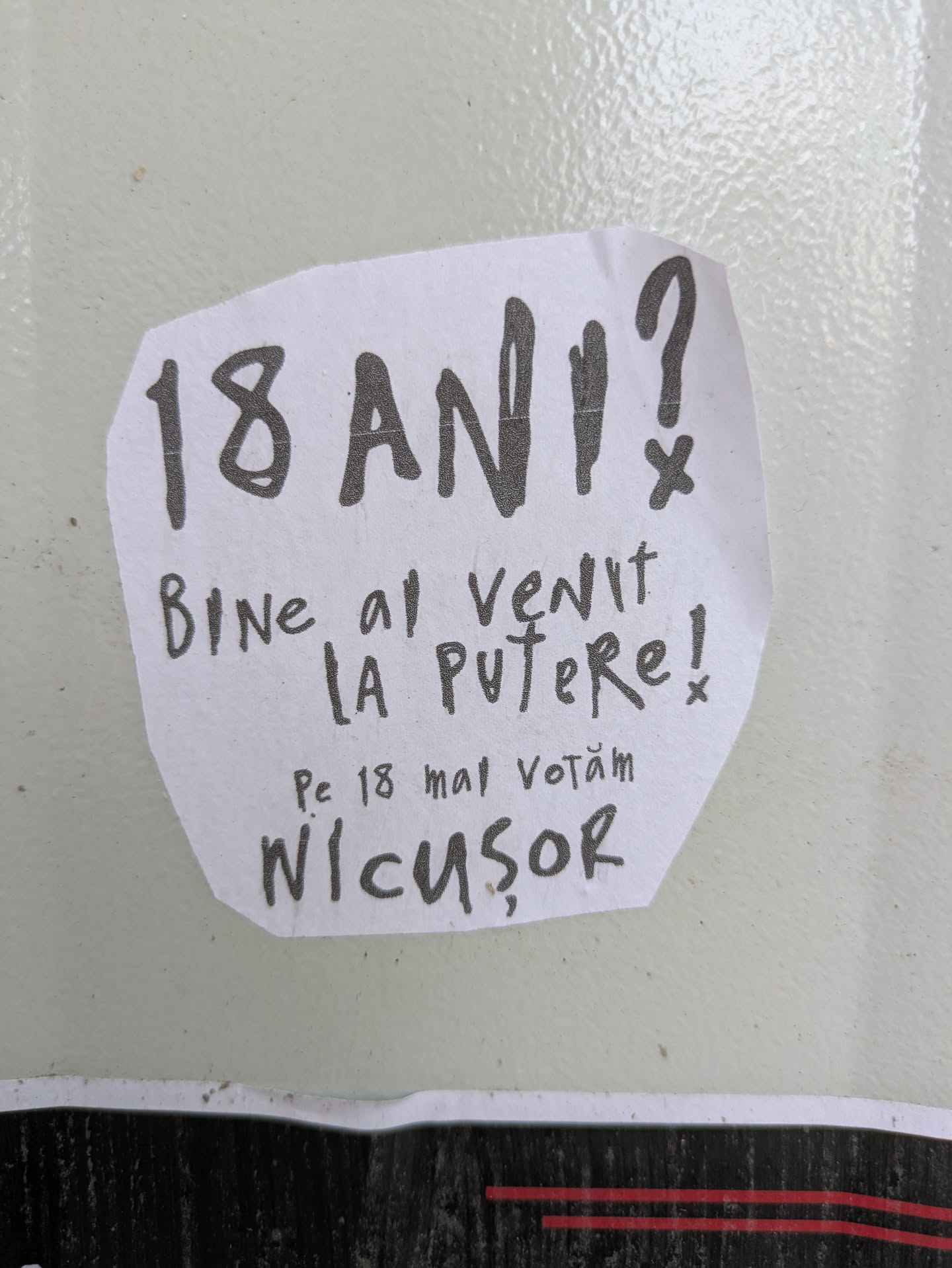 The image shows a hand-written sticker with a political message in Romanian, featuring the name 'Nicușor'. The sticker appears to be promoting a candidate, as it mentions voting on May 18th. The overall aesthetic suggests a grassroots or DIY campaign effort.