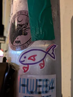 A small sticker of a stylized fish with purple and pink outlines on a white background. The fish has a simple, cartoonish design and is shown swimming to the right.  A social media handle is visible on the sticker.