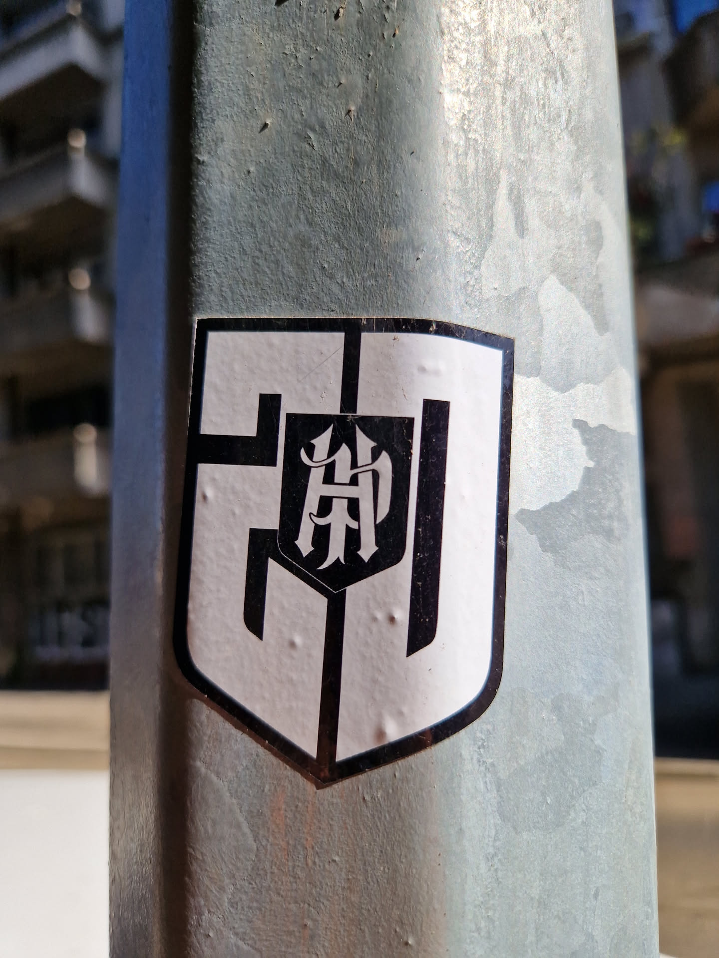 A black and white sticker in the shape of a shield. The sticker features a stylized logo with the letters "HP" in a gothic or medieval-inspired font within the shield's center. The outer edges of the sticker show a bold black outline against a white background. The sticker is affixed to a weathered metal pole.