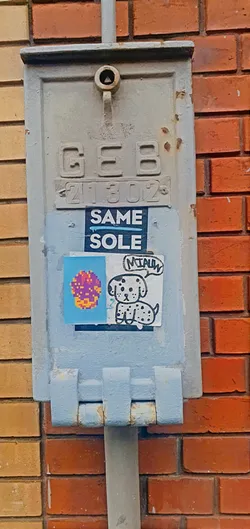 Two small stickers are attached to a gray electrical box on a brick wall. The first sticker features a pixelated mushroom in shades of orange, pink, and purple on a blue background. The second sticker shows a cartoon-style Dalmatian with spots, with a speech bubble above its head containing the word "Miau".