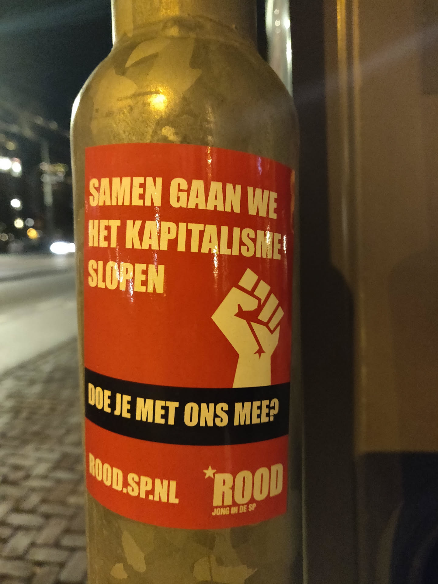 A sticker on a pole with a red background and black text. The text says "Samen gaan we het kapitalisme slopen" and "Doe je met ons mee?" with a fist in the middle and the website rood.sp.nl and the logo of Jong in de SP at the bottom.  The text translates to "Together we will dismantle capitalism. Will you join us?"