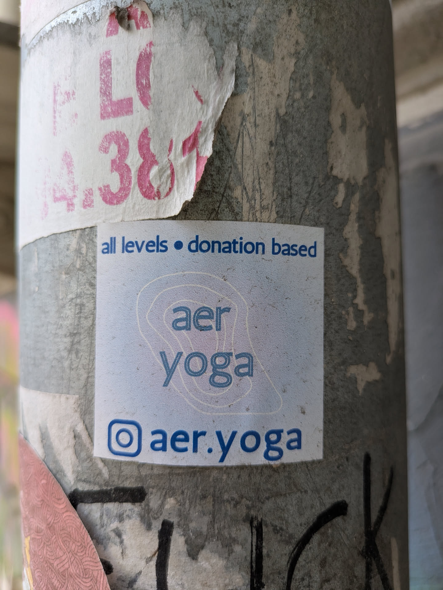 A square sticker for AER Yoga is adhered to a pole. The sticker reads "all levels • donation based aer yoga @aer.yoga" followed by the instagram logo.