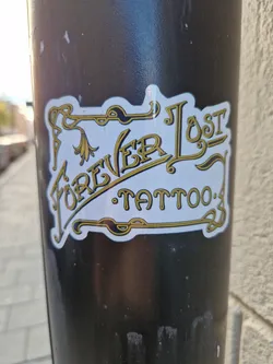 A sticker featuring the logo for Forever Lost Tattoo. The logo is in gold lettering on a white background with black ornate details. 