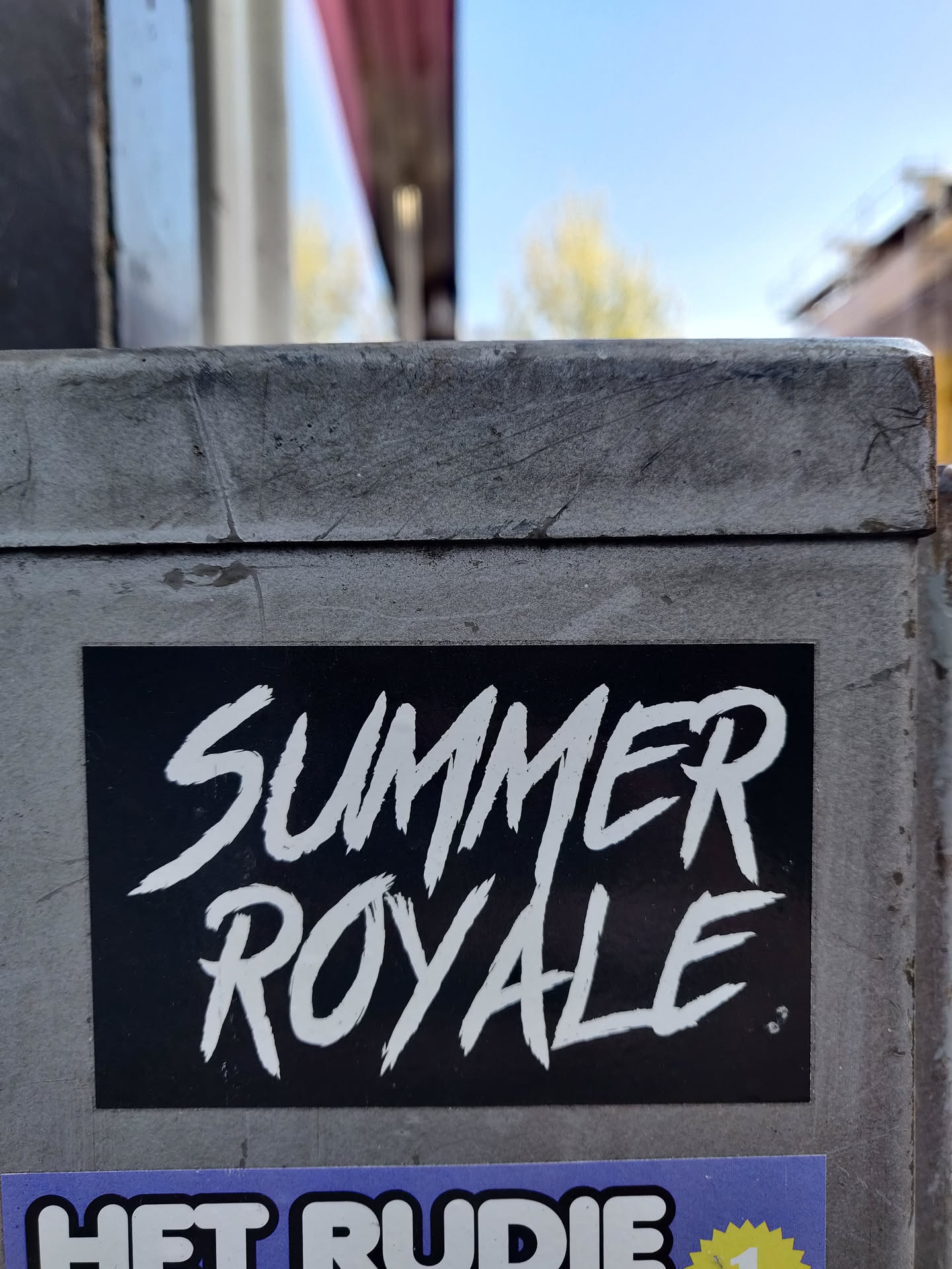 Black and white sticker with the text "Summer Royale" written in a stylized font. The sticker is affixed to a gray surface outdoors.