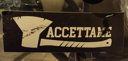 Black and white sticker with an axe and the word ACCETTAME (Italian for "Accept me") printed on it. The sticker shows some signs of wear.