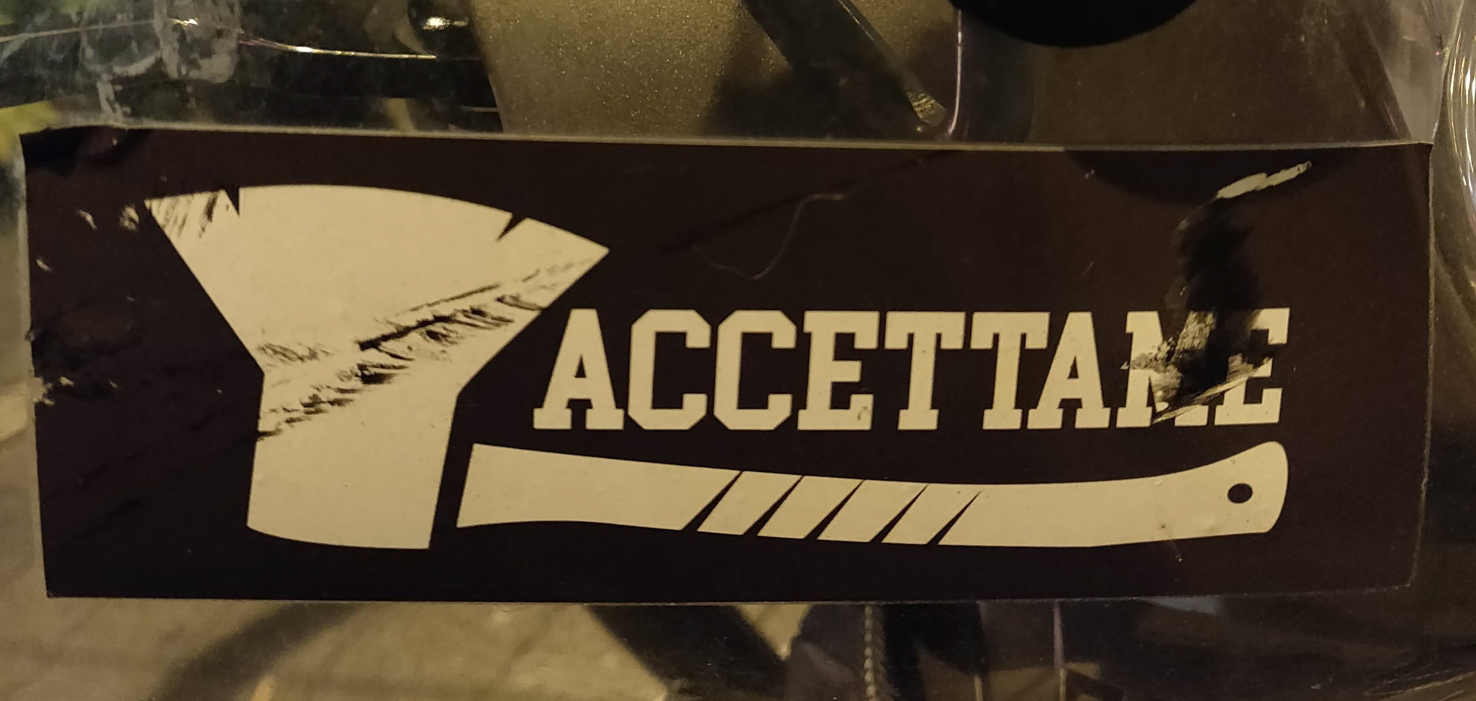 Black and white sticker with an axe and the word ACCETTAME (Italian for "Accept me") printed on it. The sticker shows some signs of wear.