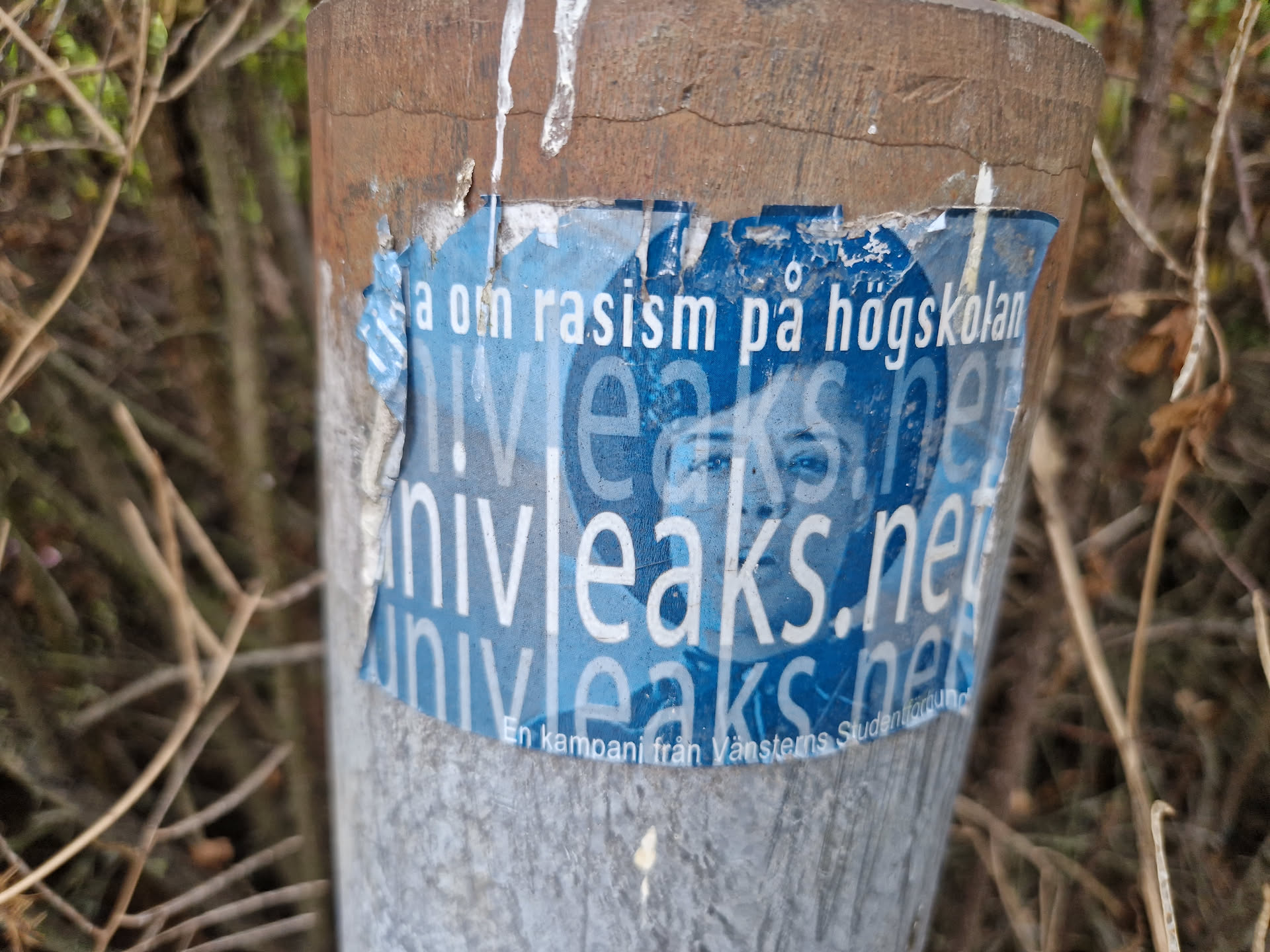 A worn blue sticker affixed to a post. The sticker features the website univleaks.net and text that translates to "Campaign about racism at the university." A faded image of a person is also present. The sticker shows signs of wear and tear.