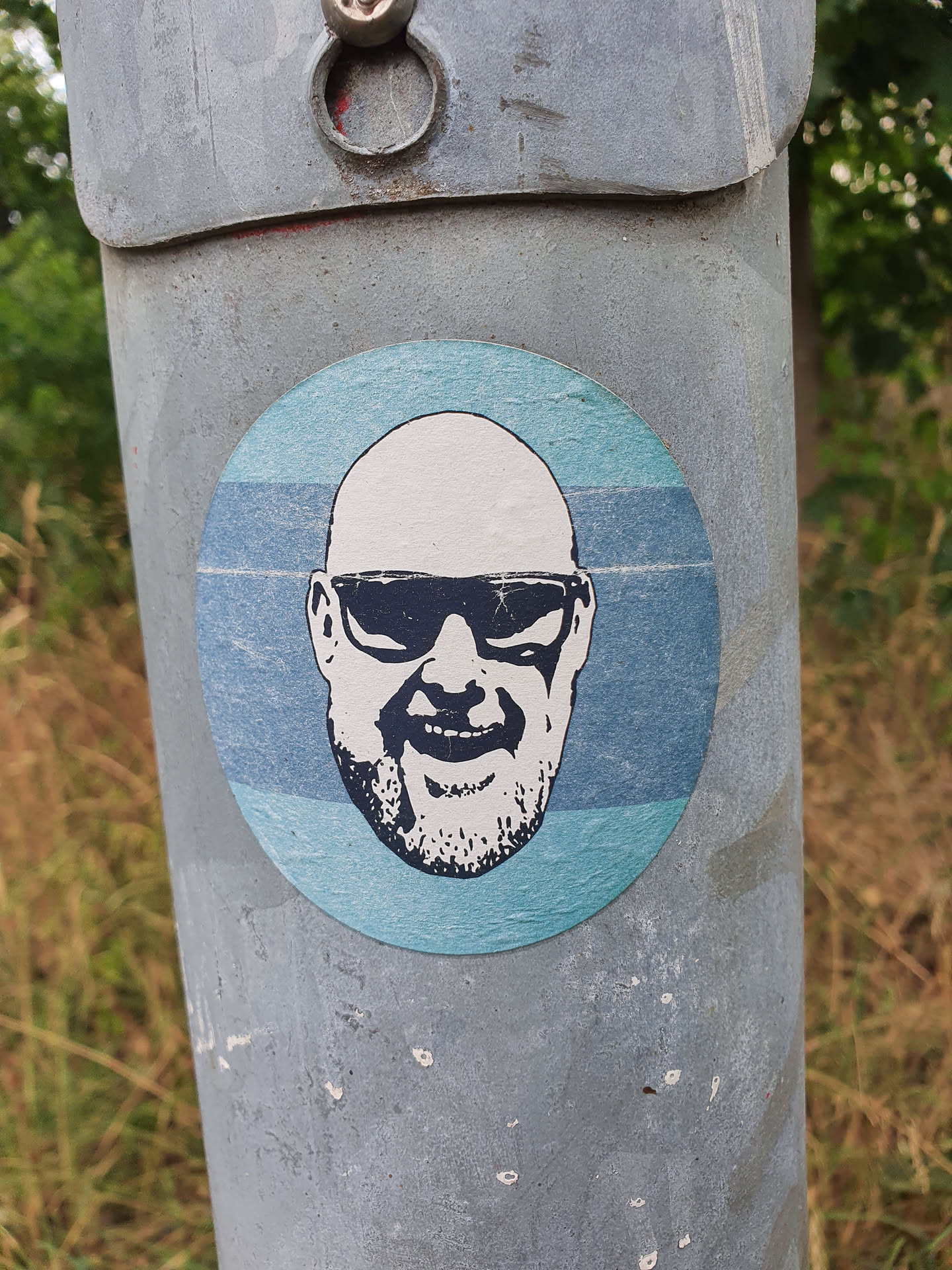 Round sticker featuring a minimalist portrait of a bald man wearing sunglasses. The background is a light blue gradient. The style is simple and effective.