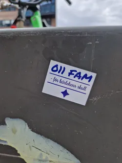 A small white sticker with blue text. It says "011 FAM - för kärlekens skull" which translates from Swedish to "011 FAM - for the sake of love". There is a small blue diamond shape at the bottom of the sticker.