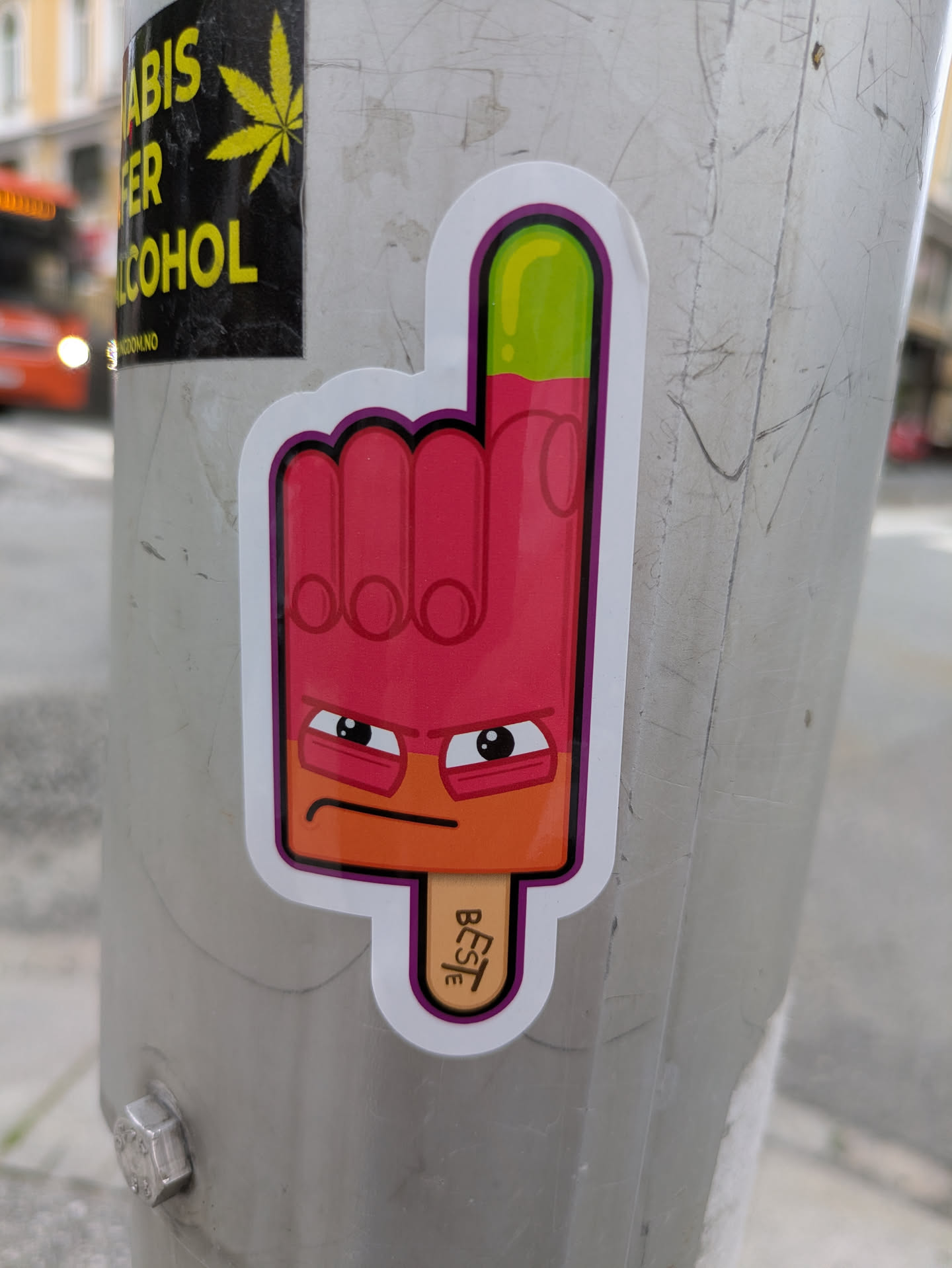 A vibrant sticker featuring an anthropomorphic ice cream popsicle shaped like a hand with the index finger raised. The popsicle is primarily bright pink/red with an orange lower section forming a face, and a lime green tip on the raised finger. It has a distinctive grumpy or determined expression with thick eyebrows and wide eyes. The sticker has a white border with a purple inner outline. A wooden popsicle stick protrudes from the bottom.