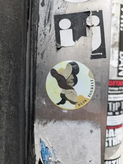 A round sticker on a metal pole features an abstract design. It includes curved shapes in yellow, brown, and black, arranged around the side profile of a woman. The sticker has the words "TOURIST" above the woman's face and "RUIN" below. The design gives off an artistic, collage-like feel.