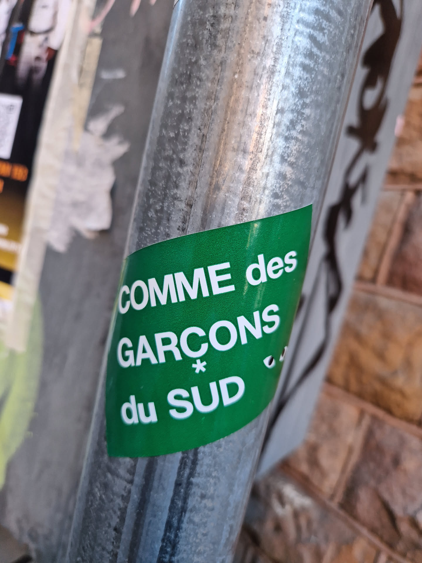 A green sticker with Comme des Garçons du Sud written on it is affixed to a metal pole. The sticker has a simple design with white text on a green background.